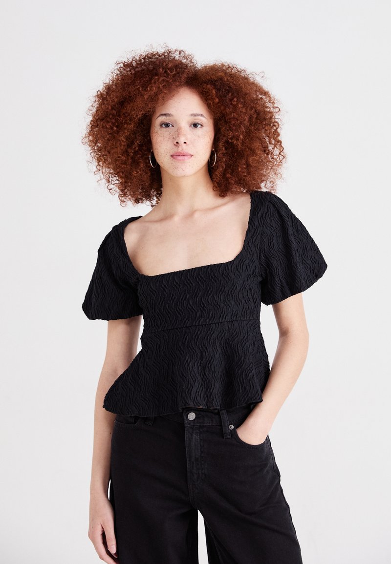 Top nero strutturato con scollatura quadrata e maniche a sbuffo, caratterizzato da un design a peplo e un delicato motivo a onde. Indossato con pantaloni scuri.