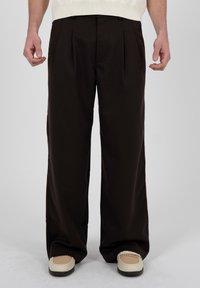 Homme portant un pantalon noir à jambes larges et des chaussures crème avec des dessus tissés beiges, se tenant devant un fond uni gris clair.