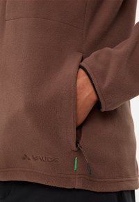 Giacca in pile marrone con una texture morbida, tasca laterale con zip e logo ricamato. Presenta rifiniture nere lungo i polsini e l'orlo.