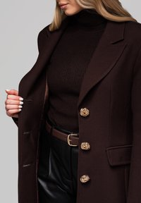 Brun uldfrakke med guld drage-mønstrede knapper, båret over en mørk ribbet turtleneck, suppleret med et brunt bælte og sorte bukser.