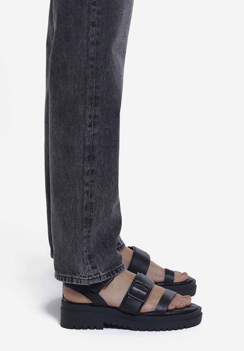 Pieds portant des sandales à plateforme épaisses noires avec des brides à boucle, associées à un jean denim gris foncé.