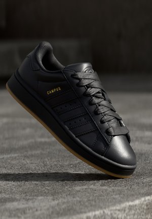 Sort læder sneaker med sorte snørebånd og gummisål, der viser siden med perforerede striber og gul tekst ""CAMPUS"".
