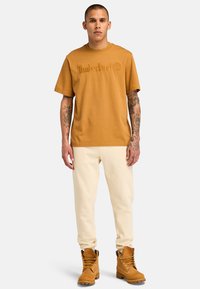Bruine katoenen T-shirt met reliëf "Timberland"-logo, off-white sweatpants en tan veterschoenen met een robuuste zool, staand tegen een neutrale achtergrond.