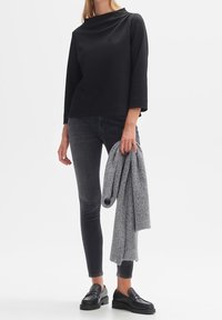 Haut noir à manches longues texturé associé à un jean foncé. Pull gris tenu dans une main, porté avec des mocassins noirs. Design simple et contemporain.