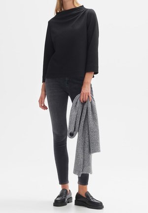 Haut noir à manches longues texturé associé à un jean foncé. Pull gris tenu dans une main, porté avec des mocassins noirs. Design simple et contemporain.