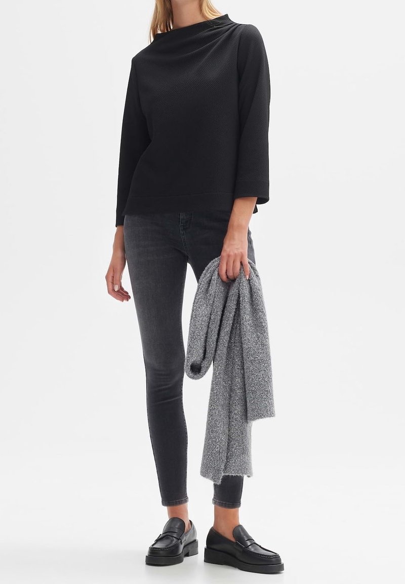 Haut noir à manches longues texturé associé à un jean foncé. Pull gris tenu dans une main, porté avec des mocassins noirs. Design simple et contemporain.