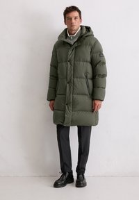 Olijfgroene donzen jas met capuchon, voorzien van een knoopsluiting aan de voorkant, ritsen aan de zijkanten en een doorgestikt ontwerp. Gecombineerd met donkere broek en zwarte laarzen.