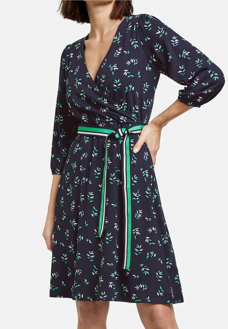 Robe portefeuille marine à manches trois quarts, avec un motif floral en vert, rose et blanc. Comprend une ceinture assortie en tissu avec des accents verts.