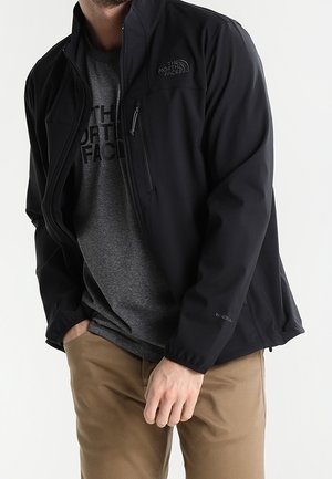 Mężczyzna w czarnej softshellowej kurtce North Face na szarej koszulce North Face i jasnobrązowych spodniach, stoi z rękami blisko kieszeni.