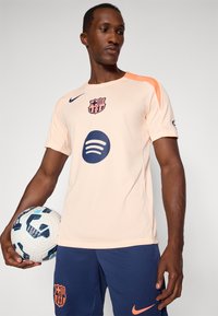 Perziken voetbalshirt met oranje schouderaccenten, voorzien van een blauw Spotify-logo en het Barcelona-embleem. Donkerblauwe shorts, met een voetbal in de hand.