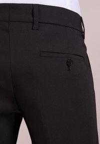 Svarte dressbukser laget av glatt stoff har flat front, beltehemper og en baklomme med knappeåpning.