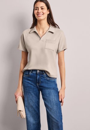 Femme portant une chemise beige à manches courtes avec col et poche poitrine, jean bleu, et tenant une pochette tissée beige sur un fond uni.