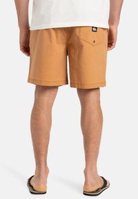 Shorts marrón claro hechos de algodón, con un bolsillo trasero con cierre de botón y un dobladillo recto. Llevados con chanclas negras.