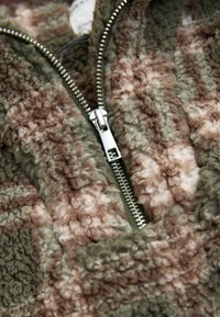 Jachetă din fleece cu fermoar, în nuanțe de verde stins, maro și crem. Are un fermoar metalic și un material moale, texturat, cu un model în dungi.