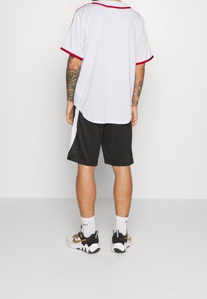 Camiseta atlética blanca con ribetes rojos, combinada con pantalones cortos negros. Mangas cortas, corte relajado. Zapatillas con detalles en negro, blanco y dorado.