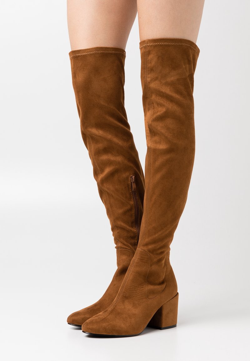 Botas de ante marrón por encima de la rodilla con tacones cuadrados y cremallera lateral, llevadas sobre piernas desnudas contra un fondo blanco.