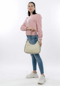 Beige läderhubbväska med en texturerad finish, svart kant och ett enda axelband. Modellen bär en rosa sweatshirt och blå jeans. Vita sneakers.