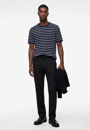 Uomo con camicia a maniche corte a righe blu navy e bianche, pantaloni neri, scarpe nere, che tiene una giacca nera nella mano sinistra.