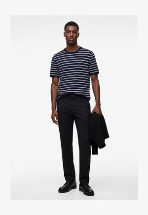 Man draagt een marineblauw-wit gestreept shirt met korte mouwen, zwarte broek, zwarte schoenen, en houdt een zwart jasje in zijn linkerhand.