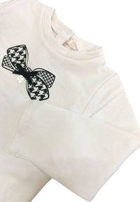 Camicia bianca a maniche lunghe con un fiocco a scacchi bianco e nero e un piccolo ciondolo decorativo. Tessuto morbido e scollatura rotonda.