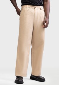 Pantalones de pierna ancha beige con una textura suave, pliegues frontales y cierre de botón, combinados con zapatos negros, de pie frente a un fondo neutro.