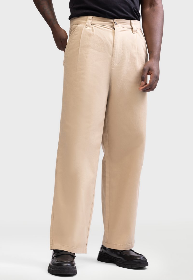 Pantalones de pierna ancha beige con una textura suave, pliegues frontales y cierre de botón, combinados con zapatos negros, de pie frente a un fondo neutro.