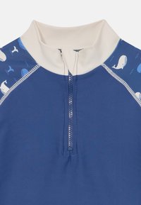Blå barnerashguard med hvit mock neck og glidelås foran. Utsmykket med et hvalmønster på kragen og skuldrene. Glatt tekstur.