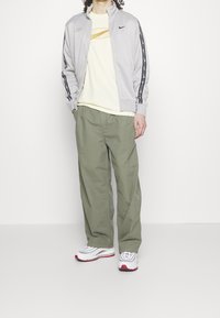 Chaqueta gris con cremallera y franjas laterales negras, camiseta amarilla con diseño geométrico, pantalones de pierna ancha verde oliva y zapatillas blancas con detalles rojos.