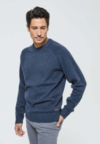 Pull en tricot bleu marine avec un col rond, des poignets et un ourlet côtelés ; présente une texture légèrement mouchetée et une petite étiquette de marque sur le côté.