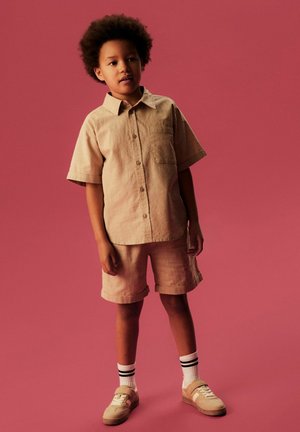 Niño con cabello rizado vestido con una camisa beige de botones de manga corta y pantalones cortos a juego, calcetines blancos con franjas negras y zapatillas beige.