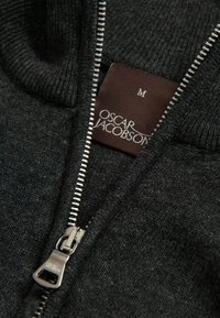 Schwarzer Reißverschluss-Pullover mit geripptem Textur, silbernem Reißverschluss und einem markierten Label mit der Aufschrift "Oscar Jacobson" vor einem dunklen Hintergrund. Größe M.