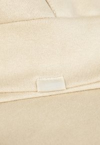 Beige gestrickter Stoff mit gerippter Textur, der im unteren Bereich ein kleines, weißes Textilschild mit schwachem Text trägt.