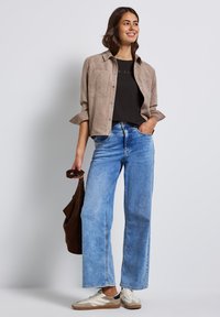 Beige suedeshirt met opgerolde mouwen, zwarte top, wijde lichtblauwe jeans en beige sneakers met bruine accenten. Bruine tas in de hand.