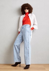 Camicia bianca oversize a bottoni, dolcevita rosso aderente, jeans blu chiaro a vita alta, stivaletti neri con tacco a spillo, e occhiali da sole scuri.
