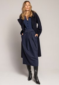 Granatowy płaszcz wełniany na bluzce w tym samym kolorze z guzikami, granatowa plisowana spódnica z wysokim stanem i czarne kozaki na obcasie sięgające do kolan.