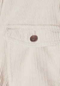 Detailní pohled na béžovou kordovou látku s klopovou kapsou uzavřenou jedním kulatým kovovým drukem.