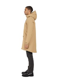 Beige vattentät parka med huva, med sidofickor och knäppta ärmavslut, i kombination med mörka jeans och svarta snörstövlar.