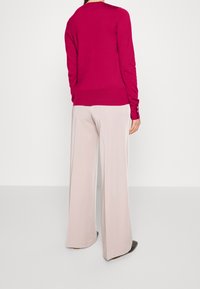 Fuchsia trui met lange mouwen, ronde hals en knoopaccenten. Gecombineerd met lichtbeige broek met wijde pijpen. Gladde textuur.