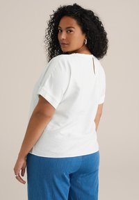 Top blanco de manga corta con escote redondeado y detalle de ojal en la espalda, hecho de una tela suave, combinado con pantalones de mezclilla azules.