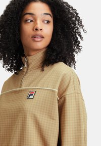 Pullover a zip da un quarto in tessuto color sabbia con un sottile motivo a quadretti, caratterizzato da un dettaglio in piping bianco e un logo multicolore sul petto.