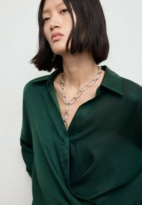 Camisa de satén verde con cuello, con un diseño drapeado. Presenta un collar de cadena plateada con formas alargadas y enlazadas. Textura suave.