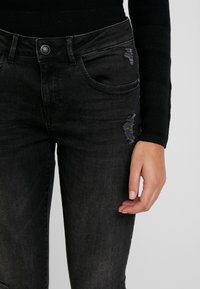 Jeans de mezclilla negra con un corte ajustado, que presentan parches desgastados, diseño clásico de cinco bolsillos y un cierre de botón metálico en la cintura.