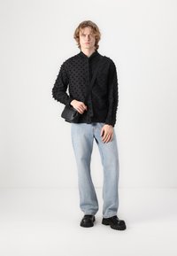 Black textured shirt with 3D dot pattern, buttoned, paired with light blue jeans and chunky black shoes, holding a small black shoulder bag.