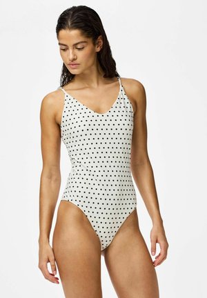 Femme aux cheveux mouillés portant un maillot de bain une pièce blanc à pois noirs, regardant vers le bas et se tenant devant un fond uni.