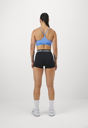 Femme athlétique debout, dos à la caméra, portant un soutien-gorge de sport Nike bleu, un short Nike Pro noir, des chaussettes blanches et des baskets blanches.