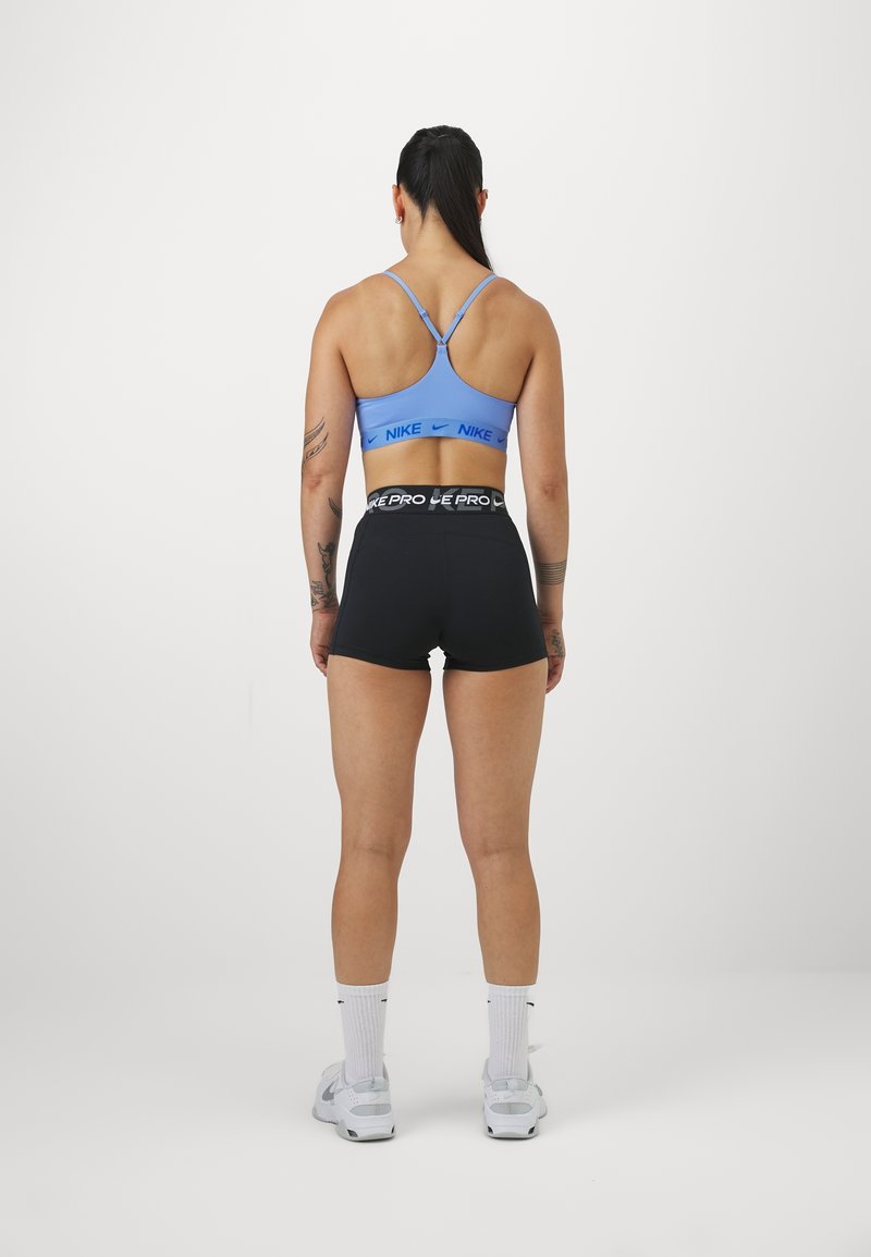 Femme athlétique debout, dos à la caméra, portant un soutien-gorge de sport Nike bleu, un short Nike Pro noir, des chaussettes blanches et des baskets blanches.