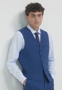 Giovane uomo con capelli ricci, che indossa un gilet a quadri blu, pantaloni abbinati, camicia azzurro chiaro e cravatta scura, appoggiato a un muro bianco.