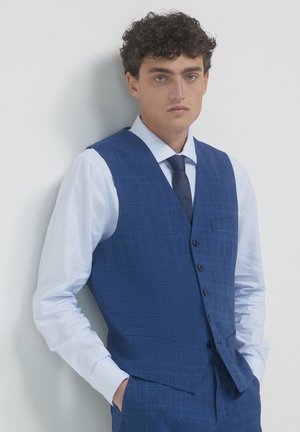 Jeune homme aux cheveux bouclés portant un gilet à carreaux bleu, un pantalon assorti, une chemise bleu clair et une cravate noire, adossé à un mur blanc.
