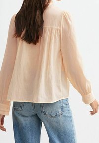 Blouse longue à manches longues de couleur pêche clair, coupe décontractée, avec empiècement plissé au dos, poignets boutonnés et tissu légèrement texturé. Associée à un jean bleu.