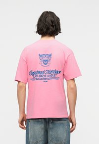 Άνδρας με κυματιστά μαλλιά που φοράει ροζ μπλουζάκι με μπλε κείμενο και έμβλημα στην πλάτη που γράφει "Εγγεγραμμένο Μέλος Laid-Back League", συνοδευόμενο από μπλε τζιν.